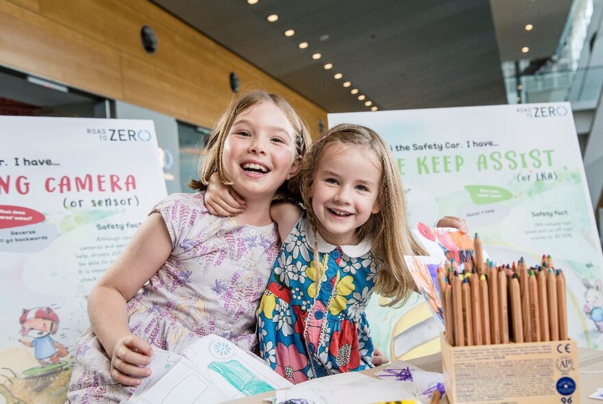 Two young girls posing with their arms around each other's shoulders, smiling, in front of two infographic boards and with a wooden box of pencils on the table in front of them.