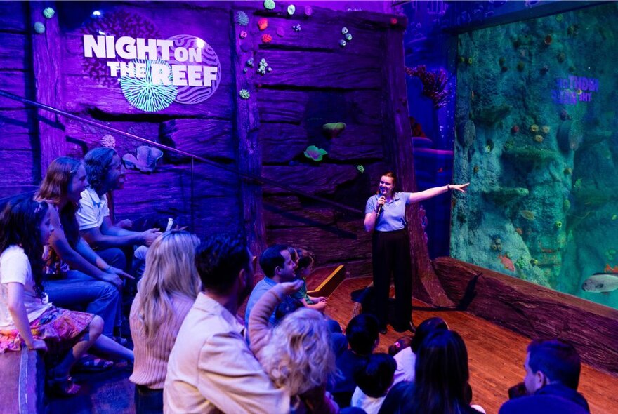 A seated audience in a purple-lit auditorium listening to a guide talk about the marine life in the aquarium behind her.