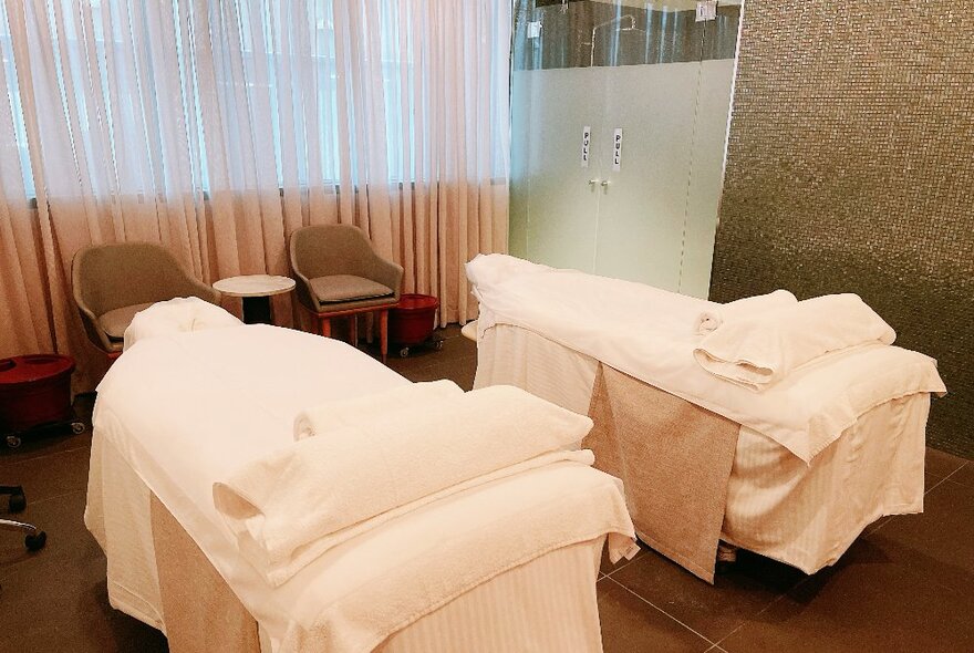 Two treatment beds in a spa treatment room, with sheer curtains over the window behind.