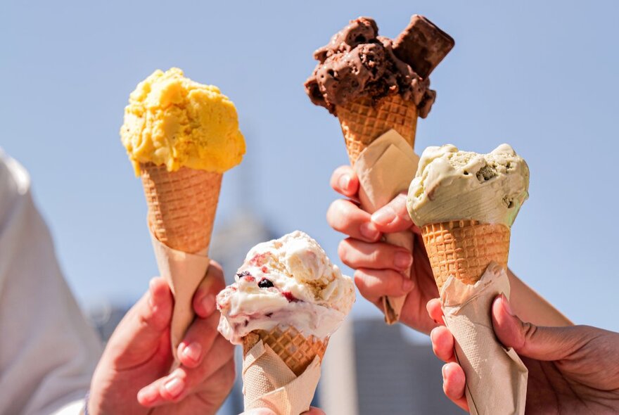 Four hands holding four ice creams in waffle cones against a blue sky.