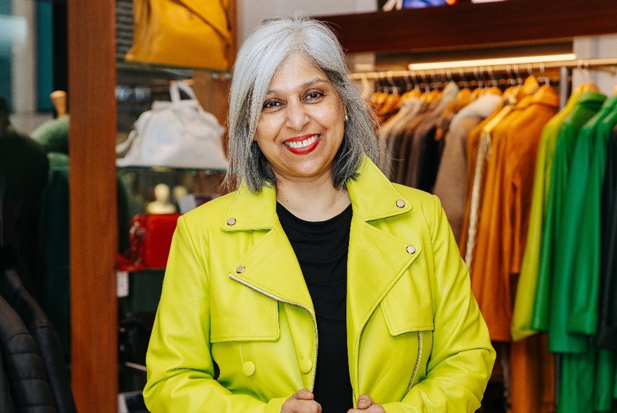 A woman with a yellow/green leather jacket in front of a colourful display of high end garments, lined up according to their colour.