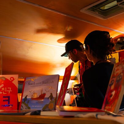 Two people inside a low-lit caravan that has multilingual books on display.
