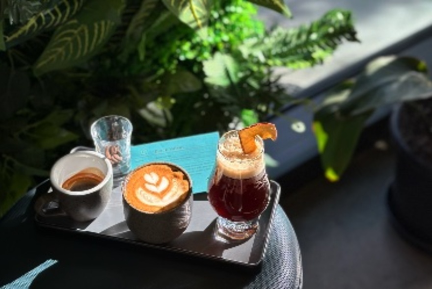 Three beverages on small tray in sun, greenery at rear.