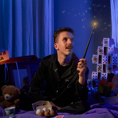 Magician, Lachlan Wilde, sitting cross legged on a cosy bed in a darkened room, wearing pyjamas and holding a wand.