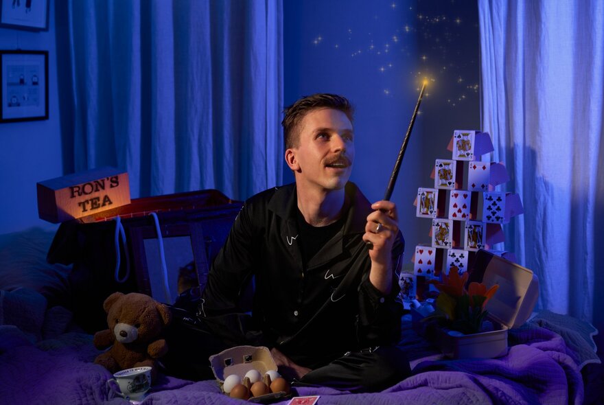 Magician, Lachlan Wilde, sitting cross legged on a cosy bed in a darkened room, wearing pyjamas and holding a wand.