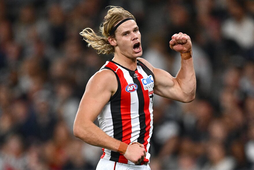A St Kilda AFL player celebrating on the field with his mouth open, both his fists clenched, a blurred stadium crowd in the background..