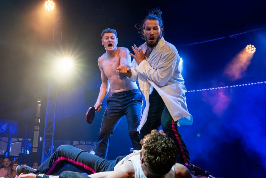 Two male performers on a brightly lit stage shout dramatically over a third person lying on the ground in the foreground.