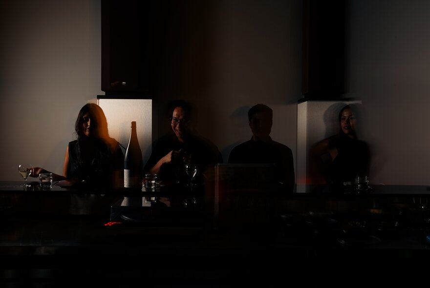 People seated in a very dark space with a bottle and glasses.