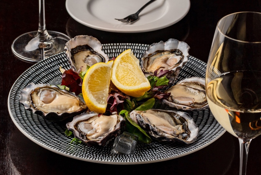 A plate of open oysters, with a lemon garnish, with a glass of wine.