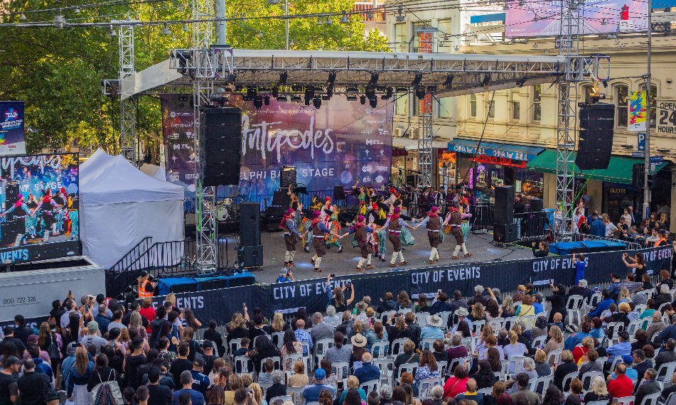 A lively outdoor street performance featuring dancers in colourful costumes on a stage with an audience seated in front.