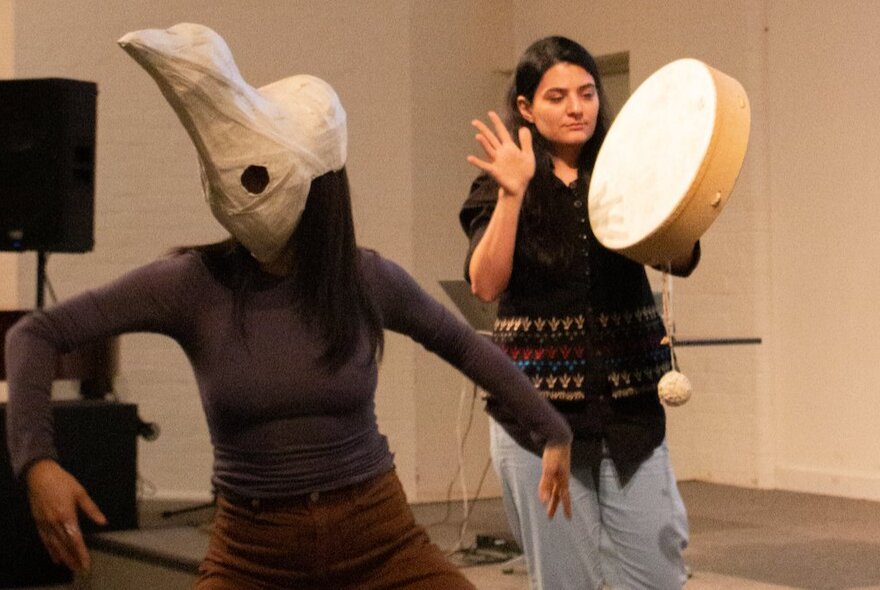 Two people performing, one of them banging a large tambourine and the other one wearing a mask and slow dancing.