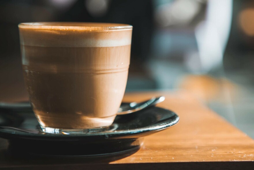 A glass of caffe latte on a black saucer with spoon, on a wooden table.