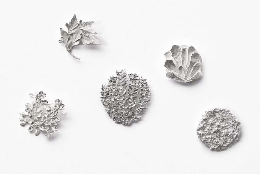 Five small, silver jewellery objects are arranged on a plain white background, each showcasing a textured or leaf-like design.