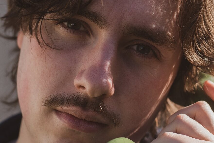 A closeup of a young man's face who is staring directly at the viewer, he holds an old phone headset to his ear.