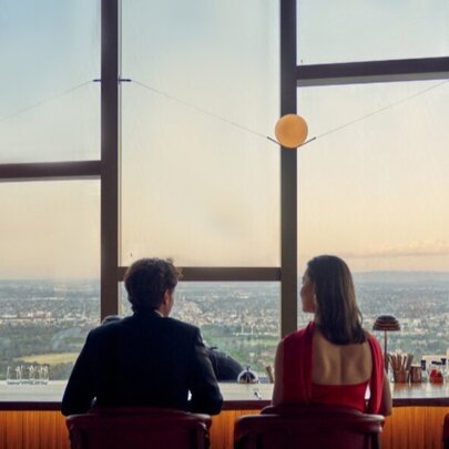 A man in a suit and a woman in a red dress sit at a bar counter, looking out a large window at a panoramic view of Melbourne at sunset.