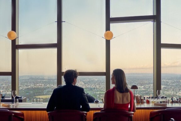 A man in a suit and a woman in a red dress sit at a bar counter, looking out a large window at a panoramic view of Melbourne at sunset.