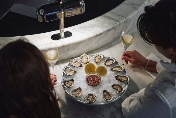 A couple seen from behind, with a plate of shucked oysters and two flutes of sparkling wine on the marble bar in front of them.