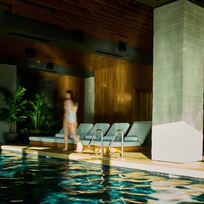 The indoor swimming pool area at 1 Hotel Melbourne, with greenery, pool lounges and a person walking near the pool's edge.