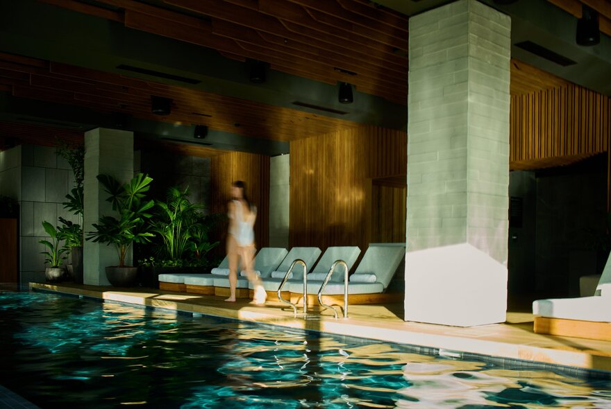 The indoor swimming pool area at 1 Hotel Melbourne, with greenery, pool lounges and a person walking near the pool's edge.