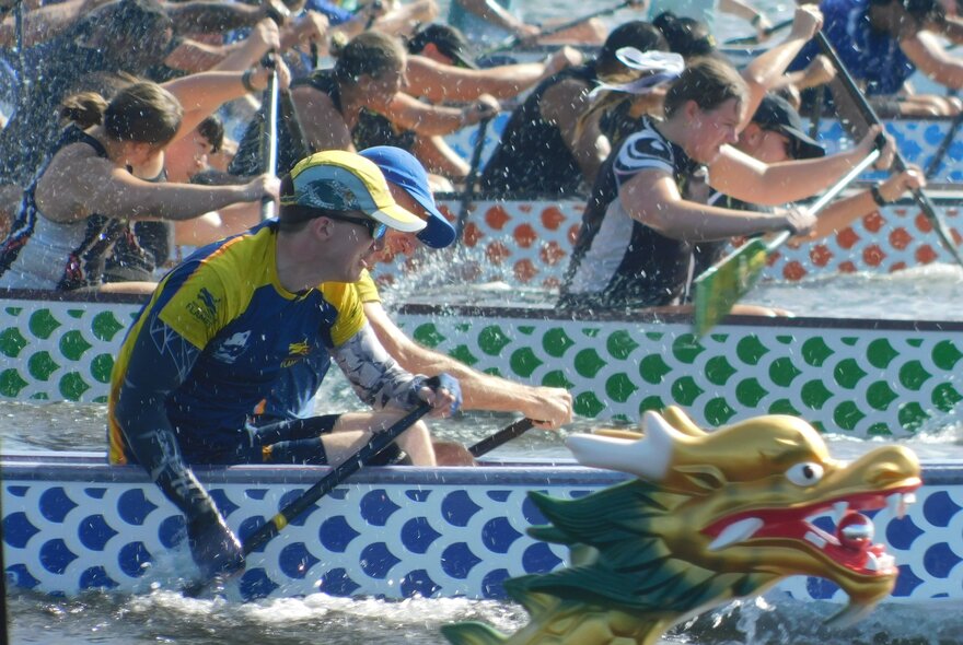 People rowing hard in a dragon boat race, the dragon head of one of the boats visible. 