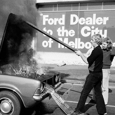An old black and white image of a man holding the bonnet of a car up as the engine burns and smoke pours out. 