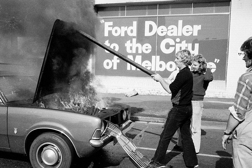 An old black and white image of a man holding the bonnet of a car up as the engine burns and smoke pours out. 