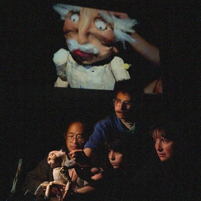 Puppeteers operating a single puppet of an old man in stage with a projection of the puppet on a large screen behind them. 