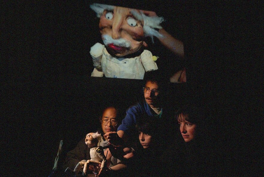 Puppeteers operating a single puppet of an old man in stage with a projection of the puppet on a large screen behind them. 