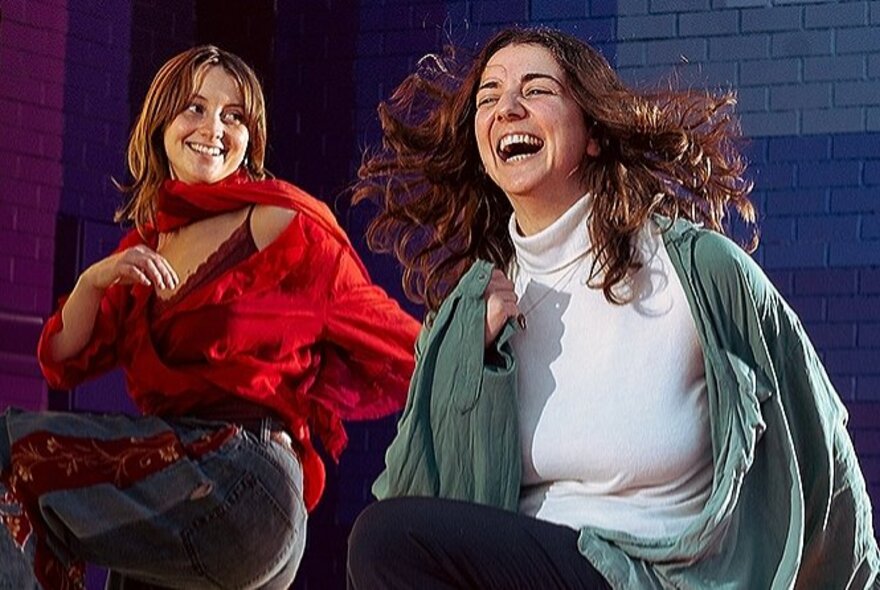 Two smiling women dancing in front of a rainbow-coloured brick wall.