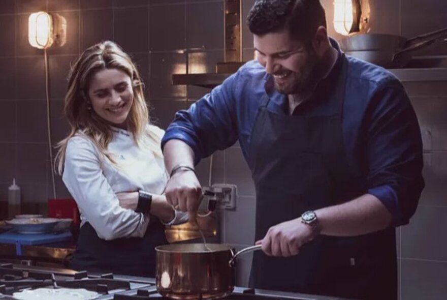 Movie still of a couple in a kitchen, the male stirring ingredients in a saucepan on a stove.
