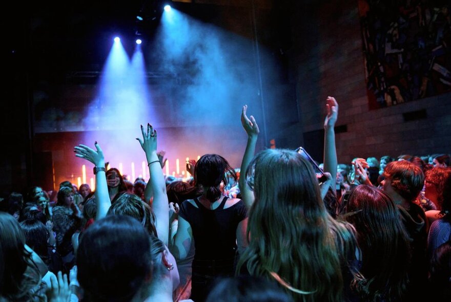 A concert with a crowd putting their hands in the air, a stage in the distance with lights and smoke. 