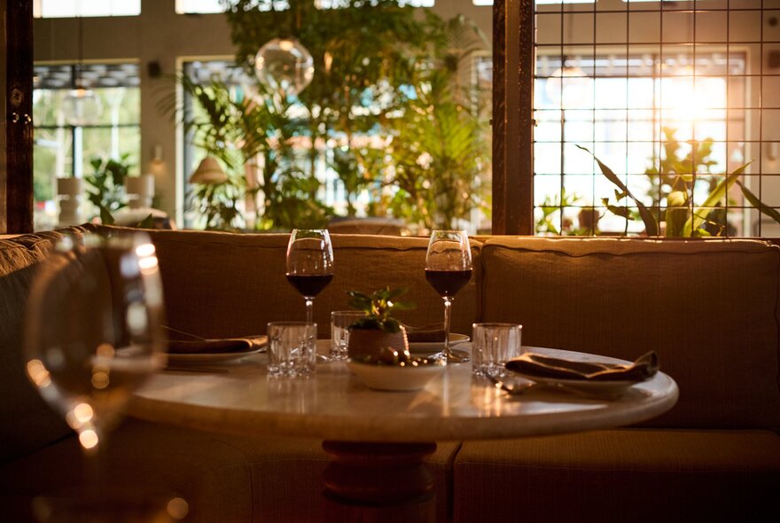 A set table in a sunlit restaurant with two glasses of red wine, windows and greenery behind.
