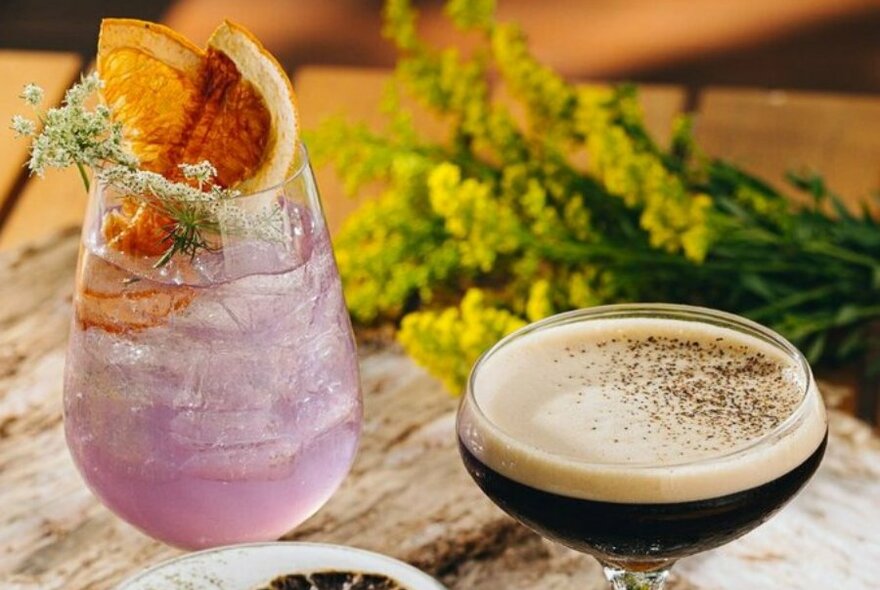 Two cocktails on wooden table, one is a lilac drink with dried fruit on top, the other an espresso martini.