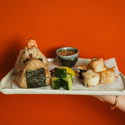 A hand holding out a plate of onigiri, diced cucumber, tempura pieces and sesame dipping sauce.