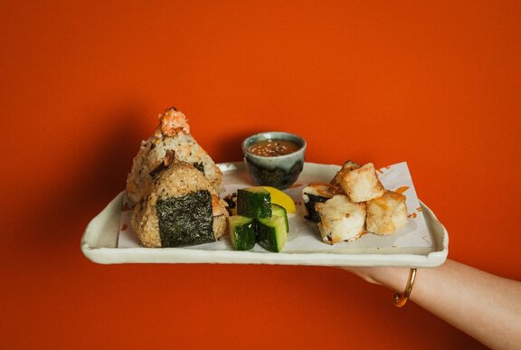 A hand holding out a plate of onigiri, diced cucumber, tempura pieces and sesame dipping sauce.