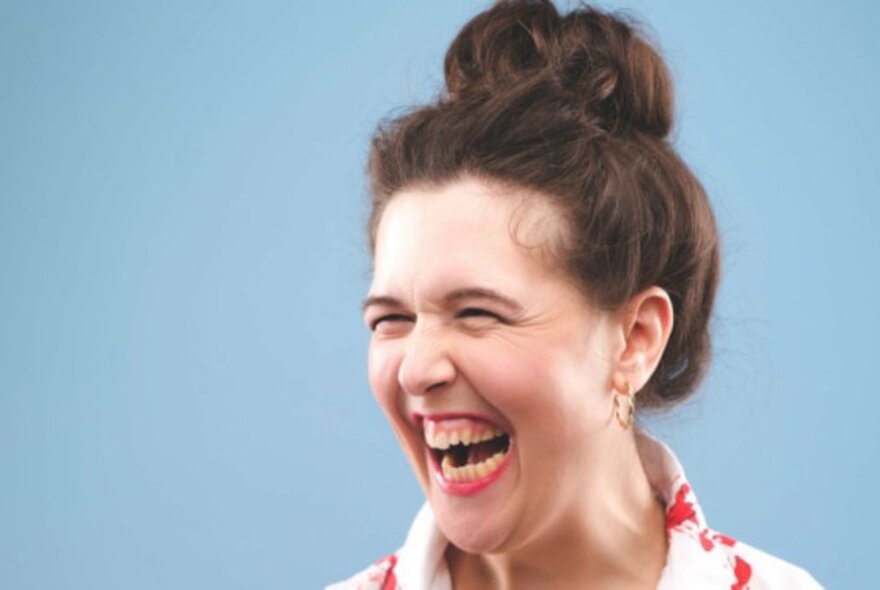 Comedian Rosie Jones with her hair in a top-knot laughing with an open mouth. 