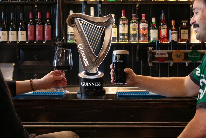 Hands holding a wine glass and glass of Guinness at a pub bar in front of the Guinness tap, with bottles lined up in the background.