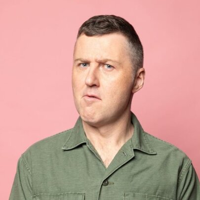 Comedian Lloyd Langford with a bemused expression, short brown hair, wearing a khaki shirt and posed against a pink background.
