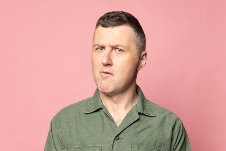 Comedian Lloyd Langford with a bemused expression, short brown hair, wearing a khaki shirt and posed against a pink background.