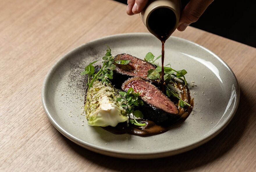 Sauce being poured from a small jug, over a steak fillet on a glazed ceramic plate.