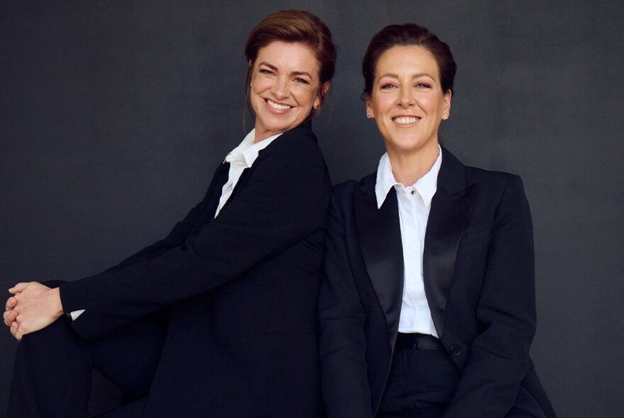 Singers Gorgi Coghlan and Anna-Lee Robertson dressed in black suits with white shirts, smiling.