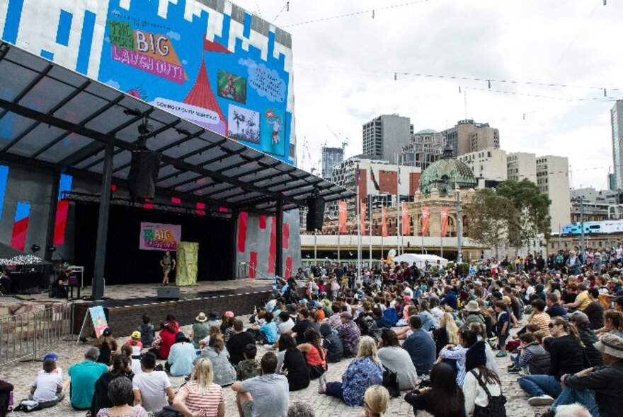 A crowd of people seated on the ground in the plaza at Fed Square enjoying comedy performances on the outdoor stage; with city buildings visible in the background.