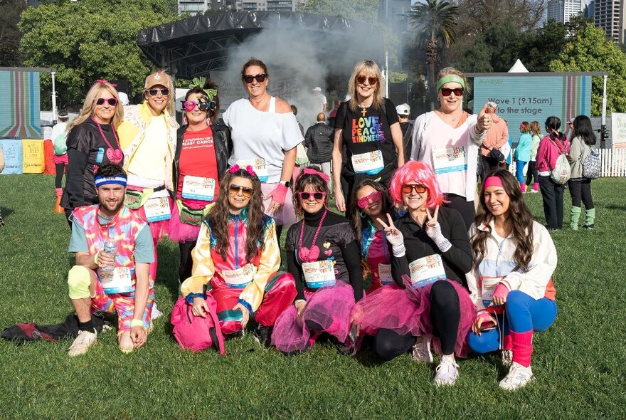 Group of people on a fundraising walk or run, dressed in bright costumes, some wearing pink wigs, posed outdoors on a lawn.