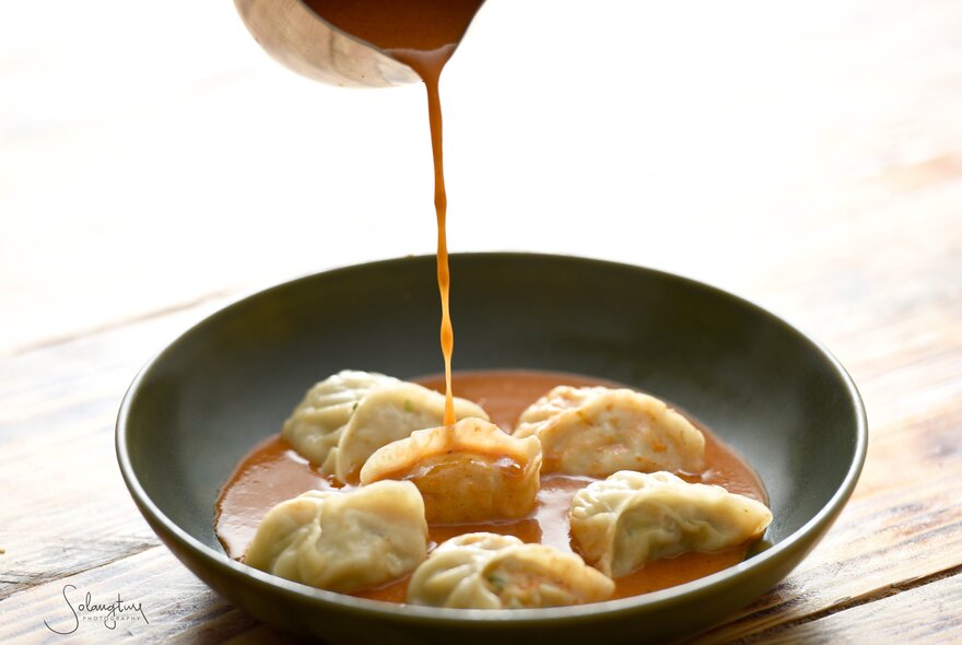 Sauce being poured from a jug into a bowl of six dumplings.