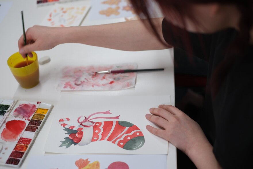 A person painting a festive image of a Christmas stocking using watercolour paint on white paper.