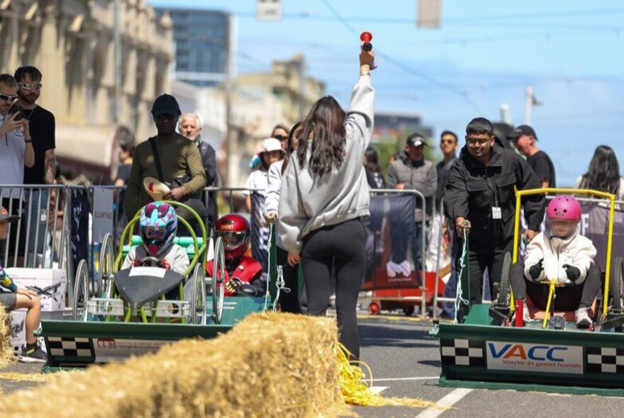 People in billy carts waiting behind a person holding a starting horn, with hay bales in the middle of the street and a few onlookers. 