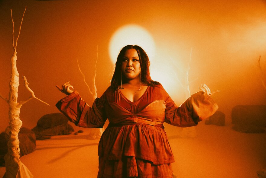 Proud Noongar woman, singer Bumpy, wearing a red and orange dress in front of an orange sky.