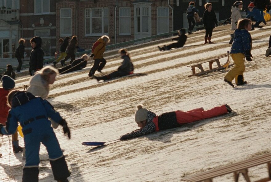 Children sliding down a hill with toboggans in the sunshine.