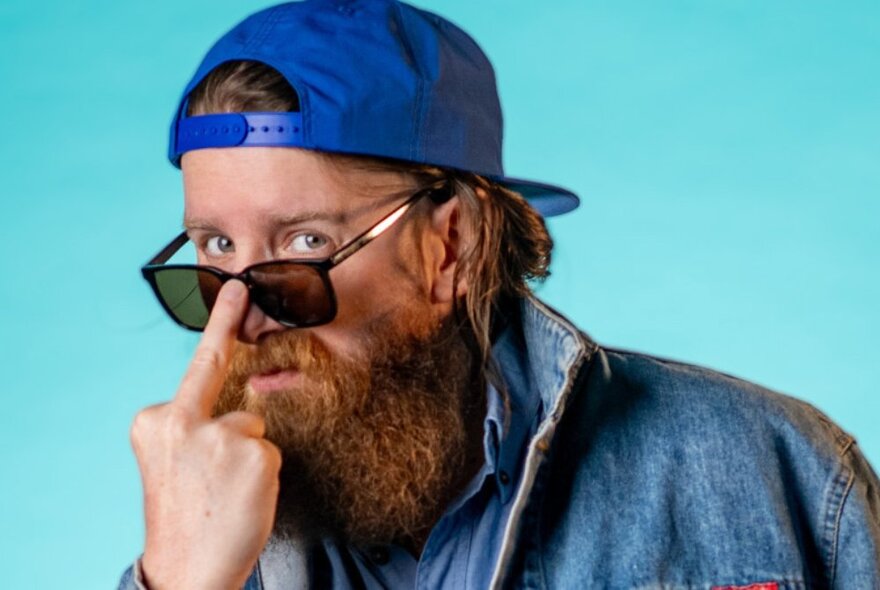 A man with a ginger beard and his blue cap on backwards, moves his sunglasses up his nose wth his middle finger in a rude gesture. 