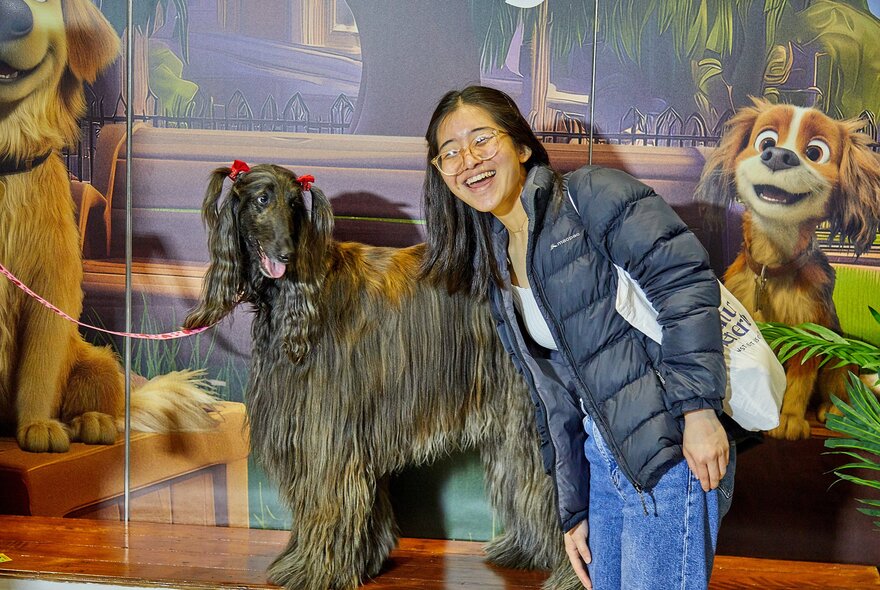 A young woman with glasses smiles broadly as she leans in next to a long-haired, dark-furred Afghan Hound wearing small red hair bows on its ears, at a dog expo.